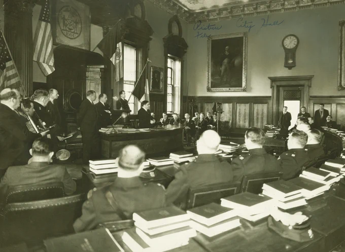 Early 20th-century courtroom scene with city officials and police personnel seated around a large table stacked with documents.