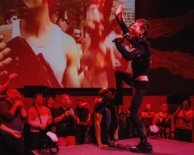 A person is performing energetically on stage with a microphone, surrounded by an enthusiastic audience. The backdrop features a large screen displaying a crowd scene. The lighting is predominantly red, creating a vibrant atmosphere.