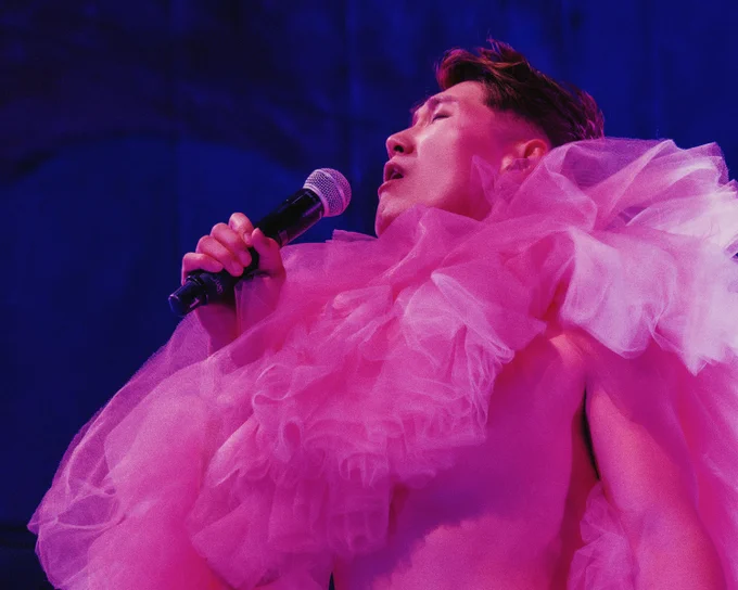 A person passionately singing into a microphone, wearing a large, fluffy pink tulle garment, against a dark blue background.
