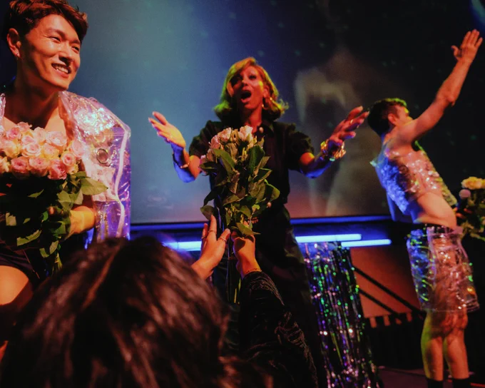 In a vibrant setting, three performers on stage joyfully interact with the audience. The central figure and one to the left receive bouquets of flowers. The background features dynamic lighting and decorative elements.