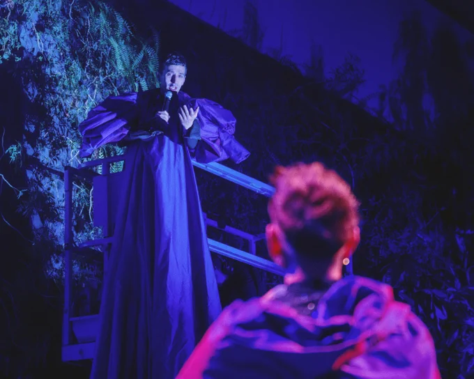 A person in dramatic, flowing attire stands elevated, speaking or performing against a backdrop of foliage. The scene is illuminated with vibrant blue and purple lighting, creating a theatrical atmosphere. Another person is visible in the foreground, listening or observing.
