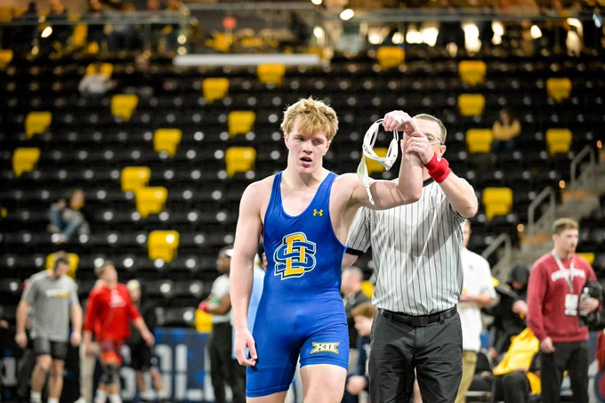 A wrestler in a blue singlet has his hand raised by a referee.