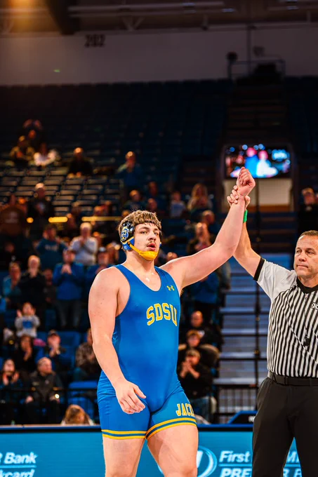 A wrestler in a blue singlet has his hand raised by a referee.