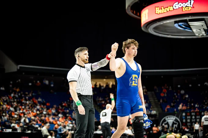 A wrestler in a blue singlet has his hand raised by a referee.