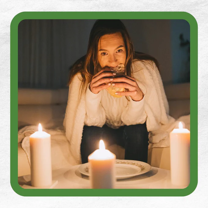 A person under a blanket eating a sandwich, surrounded by lit candles on a table.