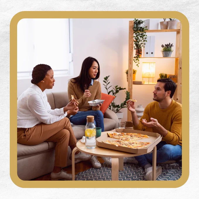 Three people in a living room eating pizza around a wooden coffee table.