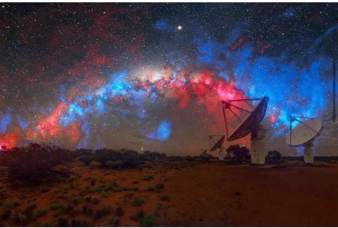 A sky filled with stars and pink and blue hues. On the right three white large parabolic antenna dishes (radio telescopes) on a barren and arid dirt landscape.