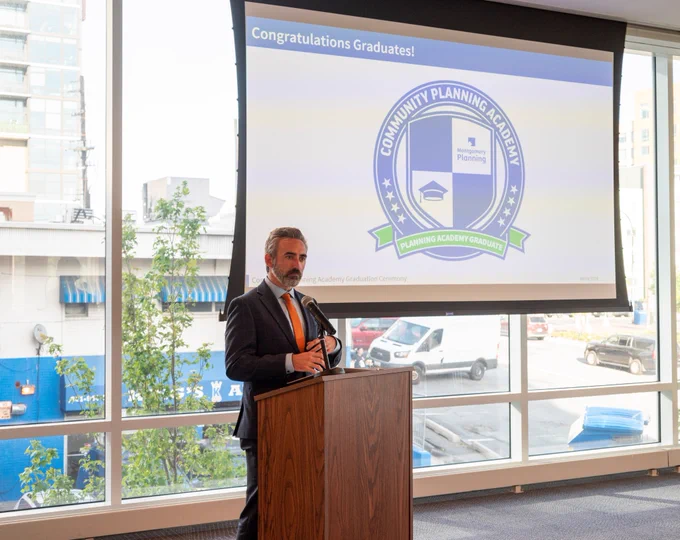A man speaks at a podium. Behind him on a screen are the words, “Congratulations Graduates!” With a round logo that reads, “Community Planning Academy.”

