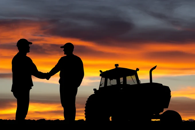 Two farmers shaking hands beside a tractor at sunset with a vibrant orange and purple sky.