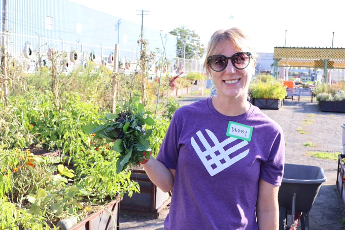 Smiling woman in sunglasses and purple volunteer shirt holding freshly picked leafy greens in a sunny community garden.