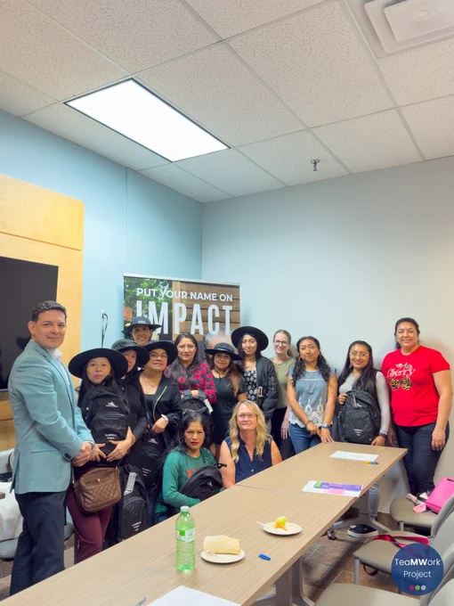Foto de grupo de los asistentes y organizadores de la presentación, reunidos en una sala de reuniones; muchos de ellos sostienen mochilas y materiales del Proyecto TeaMWork, y al fondo se ve una pancarta sobre el impacto en la comunidad.