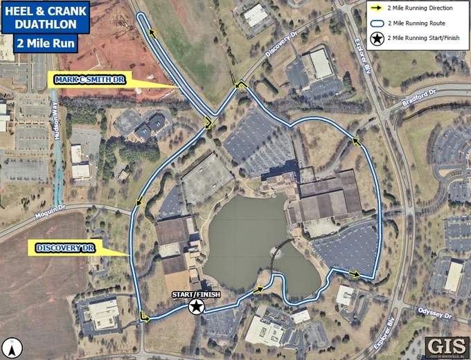 An overhead map displays the 2‑mile running route for the Heel & Crank Duathlon. A blue line outlines the running course, with yellow arrows indicating the direction of travel. The loop begins and ends at a black‑and‑white star labeled “Start/Finish,” located near a lake and several buildings. The route follows sections of Explorer Boulevard, Discovery Drive, Moquin Drive, and Mark C. Smith Drive, weaving around office buildings, parking lots, open fields, and landscaped areas. A legend in the upper right corner explains symbols for running direction, running route, and the start/finish point. Street names and landmarks are labeled throughout the map, and a GIS logo for the City of Huntsville appears in the lower right corner.