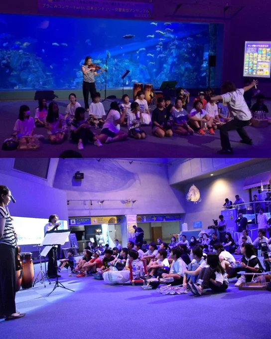 A lively group gathers in an aquarium setting with large tanks and colorful fish in the background. Some individuals sit while others stand or gesture animatedly. A microphone stand and musical instruments, including drums, are visible. Bright lights and display panels add to the engaging atmosphere.