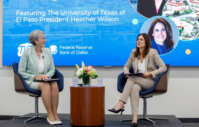 Two individuals, UTEP President Heather Wilson and Lorie Logan are seated on a stage with a backdrop featuring The University of Texas at El Paso and the Federal Reserve Bank of Dallas logos. A floral arrangement is on the small table between them. 
