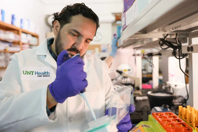 A person in a lab coat with "UNT Health Fort Worth" on it works in a laboratory using a pipette. The background shows laboratory equipment and supplies.