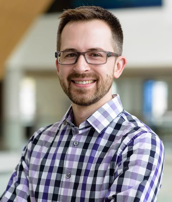 A man wearing glasses and a button down shirt is facing forward and smiling.