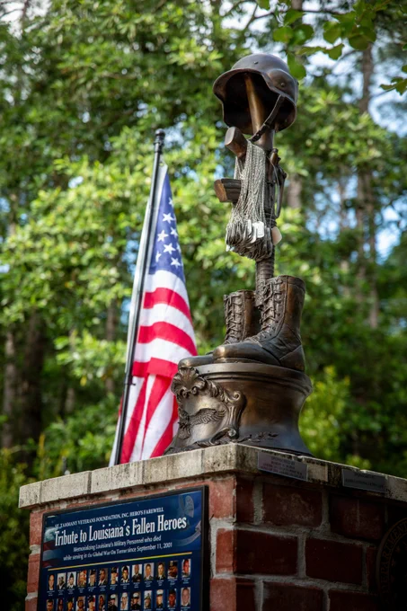 A battlefield cross memorial with combat boots, helmet, and dog tags stands beside an American flag at a tribute honoring fallen service members.