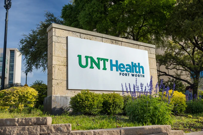 A large sign with "UNT Health Fort Worth" is prominently displayed in a landscaped area. The sign is surrounded by greenery and purple flowers, with trees in the background.
