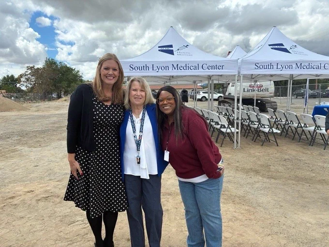 Group of three people smiling and standing outside in front of South Lyon Medical Center tents. Rows of chairs are visible in the background.