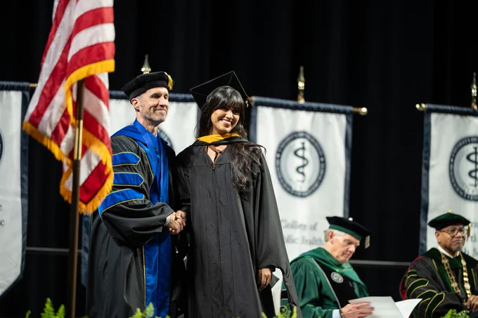 A person in academic regalia receives a diploma on stage while shaking hands with a faculty member at a graduation ceremony. The U.S. flag and banners with academic insignia are in the background. Other seated faculty are visible in the background.