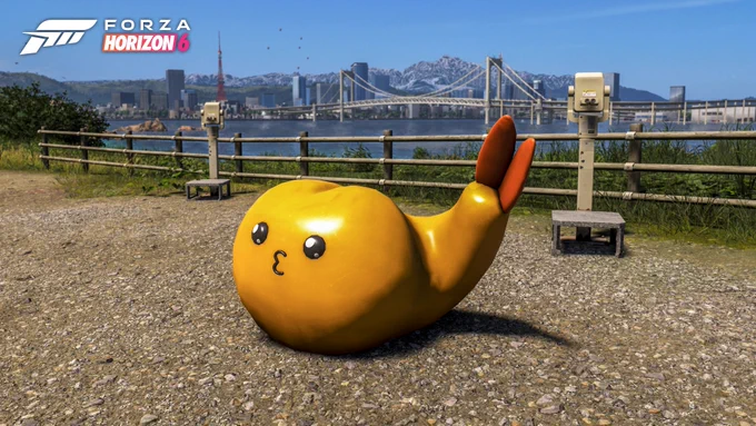 A cute, cartoonish yellow whale sculpture with orange fins is perched on a gravel surface. In the background, there's a scenic view of a bridge and a city skyline with mountains under a clear blue sky. The "Forza Horizon 6" logo is in the top left corner.