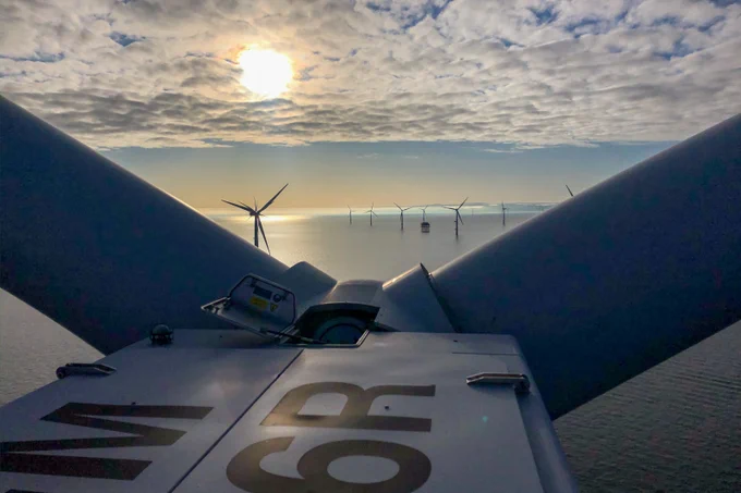 A scenic view from the top of a large wind turbine over a calm sea, with several other wind turbines visible in the distance. The sun is partially obscured by clouds, creating a serene atmosphere.