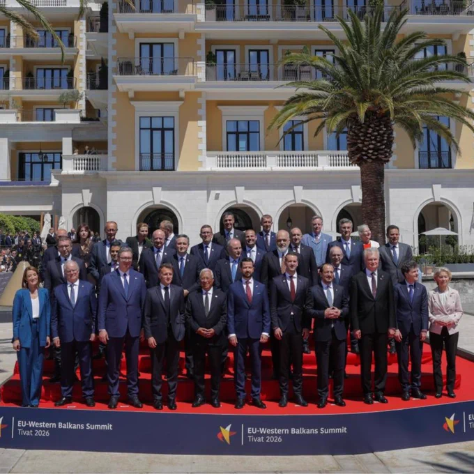 A wide, eye-level shot captures a large group of delegates in suits posing for a group photograph on a red carpet at the EU-Western Balkans Summit in Tivat, 2026. They stand in front of a grand, multi-storey yellow and white building with balconies, flanked by two large palm trees. On the right, a marina is visible with boats moored along the dock under a partly cloudy blue sky, while a crowd of spectators stands behind a barrier on the left.