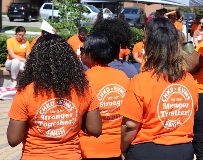 Group of people wearing bright orange shirts with 'We are Stronger Together!' printed on the back, gathered outdoors in a sunny setting.