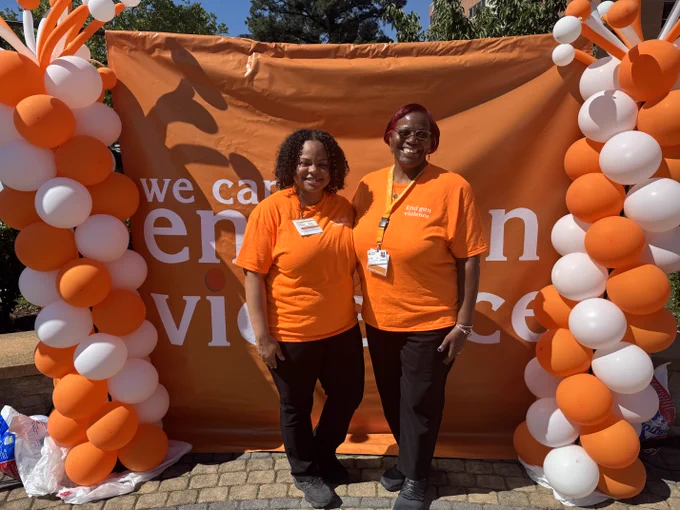 Two women wearing matching orange shirts stand in front of an orange backdrop with white text about ending violence, flanked by orange and white balloon columns.