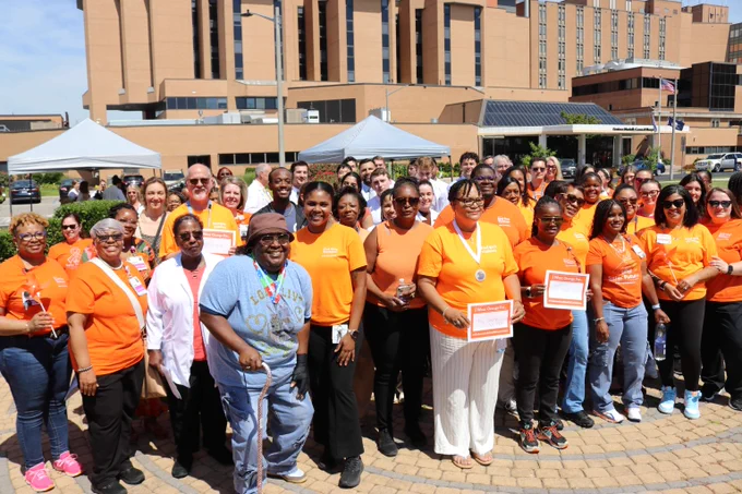 Large group of diverse people gathered outdoors, many wearing bright orange shirts, some holding certificates and medals, smiling under clear skies.