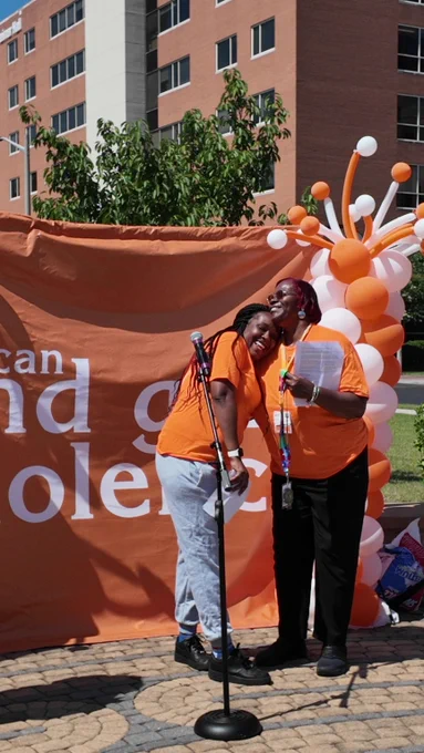 Two women in orange shirts smiling and posing together in front of an orange banner and balloon decoration outdoors.