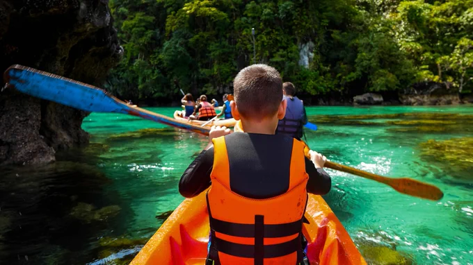 Kids and camp counselors kayak on crystal clear stream bordered by trees and a waterfall, wearing colorful life jackets. Image by Jane Foster on Pexels.