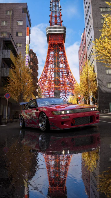 Un coche deportivo rojo está estacionado en una calle mojada con un charco reflectante. La icónica Torre de Tokio se ve claramente en el fondo, enmarcada por altos edificios y árboles de colores otoñales. El cielo está claro y azul.