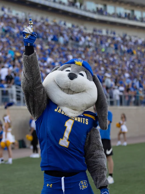 A mascot in a Jackrabbits uniform, with the number one displayed, is on a football field. The mascot is raising a finger and smiling. A crowd in blue and yellow fills the stadium in the background.