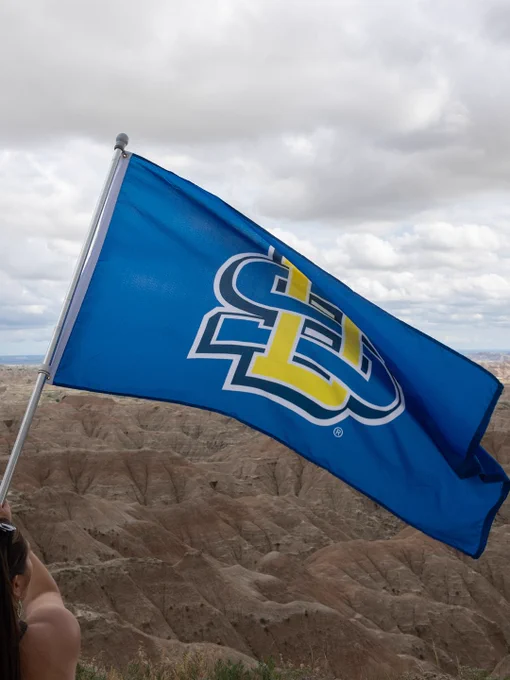A blue flag with the South Dakota State University logo is being held against a backdrop of rugged, rocky terrain under a cloudy sky.