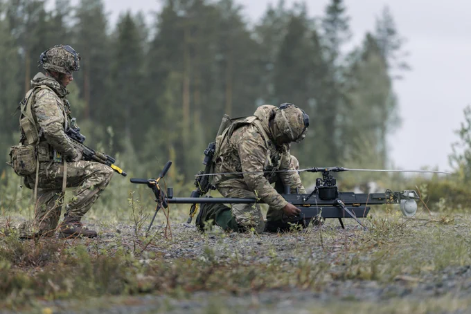Two people in camouflage military uniforms are working outdoors with drone equipment. One is kneeling beside the drone while the other stands nearby, observing. They are in a grassy area with forest in the background.