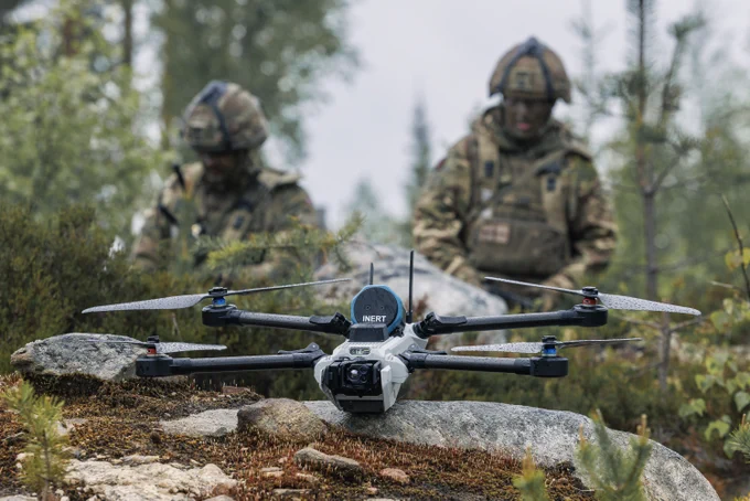 A drone by INERT sits on the ground in a forested area, with two individuals in camouflage gear and helmets standing in the background.