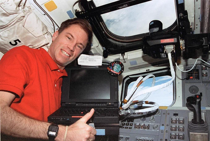 Richard Searfoss smiles widely while giving a thumbs up while posing with a laptop computer and camera. He is wearing a short-sleeved red polo shirt, and the windows of the space shuttle are visible behind him.