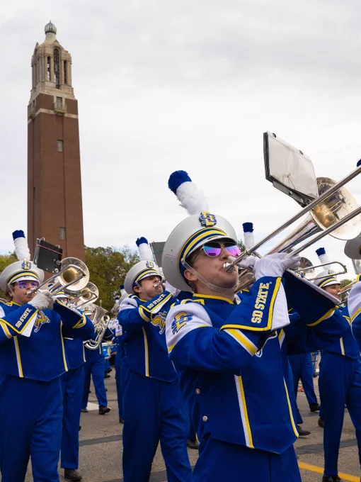 A marching band in blue and yellow uniforms plays brass instruments near a tall brick clock tower. The musicians wear hats with blue plumes and "SDSU" is visible on their uniform sleeves.