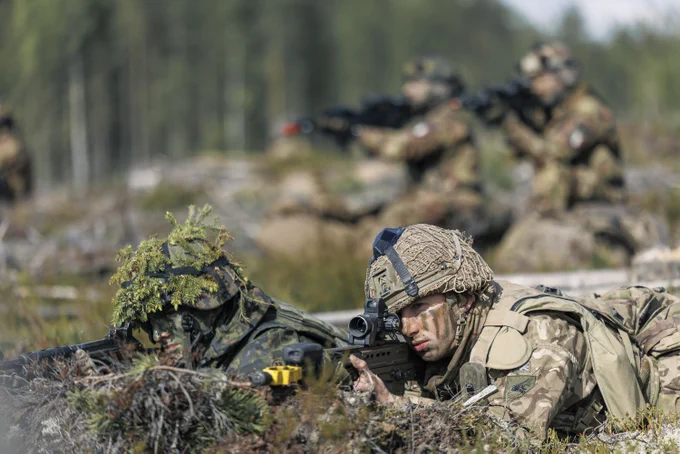 Soldiers wearing camouflage uniforms and tactical gear lie prone on the ground in a forest. They are aiming rifles, with one soldier in the foreground focusing intently through the scope. The background shows other soldiers similarly engaged, and the setting is outdoors with trees and natural debris.