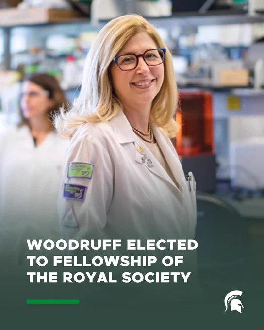 Person wearing a white lab coat and glasses stands in a laboratory setting, with blurred lab equipment and another lab-coated person in the background. Overlaid text at the bottom reads, “WOODRUFF ELECTED TO FELLOWSHIP OF THE ROYAL SOCIETY,” alongside a Michigan State University Spartan logo.