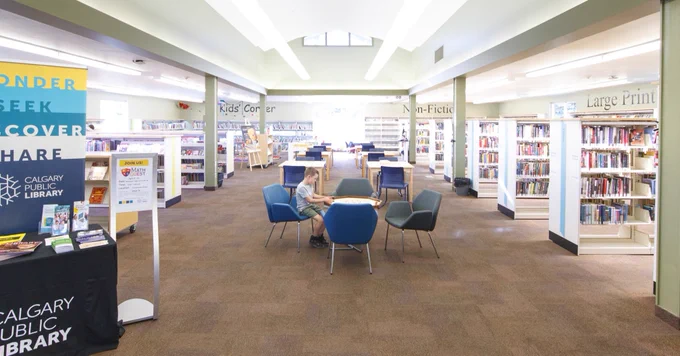 A spacious library interior with bookshelves lining the walls and several chairs arranged around a table. A Calgary Public Library sign and informational displays are visible on the left. The space is well-lit with overhead lighting.
