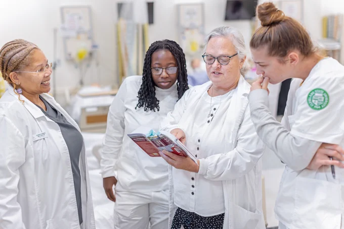 A group of CSU Nursing students dressed in scrubs reading a book with a professor in the lab. 