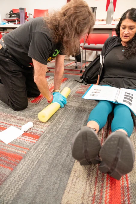 A person is kneeling on the floor, wrapping a yellow foam roller. The seated individual is holding a printed manual and is lying back on a patterned carpet. Red chairs and a table with bottles are in the background.