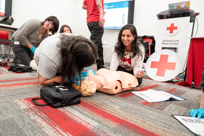 Participants are engaged in a CPR training session with an American Red Cross sign visible. One person is practicing chest compressions on a CPR manikin, while others observe and provide guidance. The setting is a classroom with instructional materials on the floor.