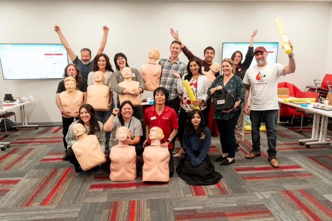 A group of people in a room standing and kneeling with CPR practice mannequins. Some participants are raising their arms joyfully. There are two screens on the wall displaying diagrams. The room has a patterned carpet and several red chairs. Some attendees are holding certificates and American Red Cross training materials.