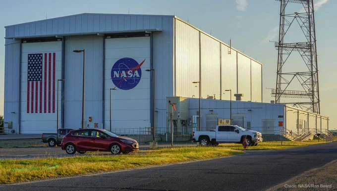 The Horizontal Integration Facility at NASA’s Wallops Flight Facility in Virginia, a large, white building with two tall doors. The left door is painted with an American flag and the right door with the blue NASA logo. A few cars are seen in the parking lot in front of the building. The image is watermarked “Credit: NASA/Ron Beard.”