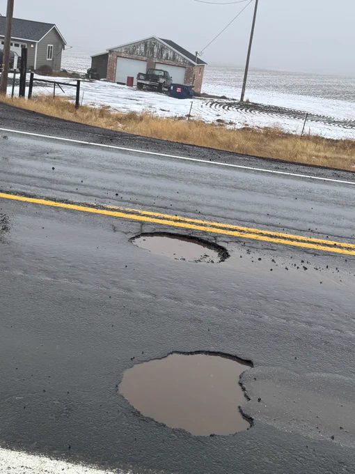A section of U.S. 97 between Moro and Grass Valley with two large potholes filled with water in the foreground. In the background, there is a house and a separate garage with a tractor parked next to it. The surrounding fields are dusted with snow.