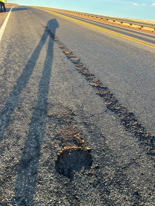 A long shadow stretches across an empty section of U.S. 97, highlighting cracks in the pavement. The horizon is slightly lit by the setting or rising sun. A guardrail runs parallel to the road on the right side.