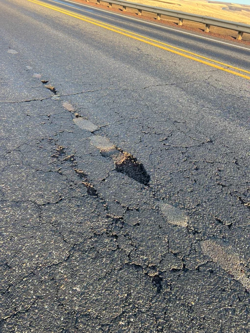 A cracked and uneven road surface with a visible pothole. This section of U.S. 97 between Moro and Grass Valley includes yellow dividing lines and a metal guardrail on the right side. The sky is clear.